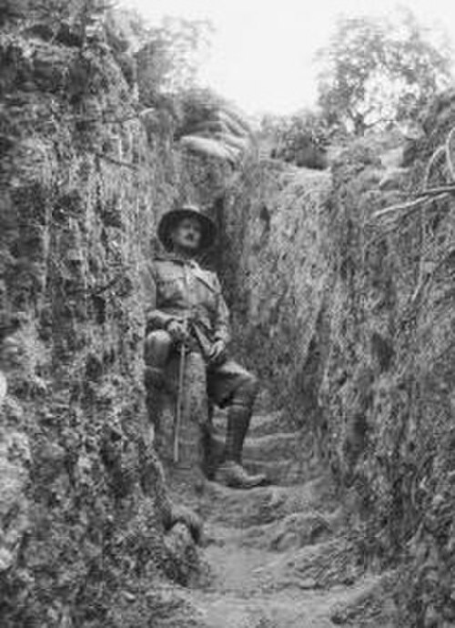 塹壕の中に座っている軍服の男。両側には土の壁があり、その上に土嚢が置かれている。