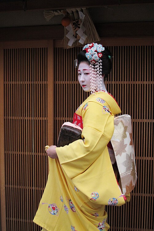 若い芸者見習いが伝統的な日本建築の外に立っていました。彼女の黄色い着物の袖は舞妓さんのものより短く、髪飾りも舞妓さんよりたくさんつけています。