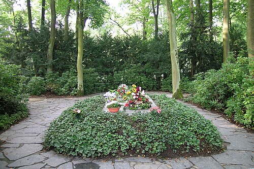 背の低い葉の植物でいっぱいの大きな花壇の真ん中にある平らな墓石の上に、いくつかの花の供え物が置かれています。花壇の両側には、不規則に舗装された小道があります。