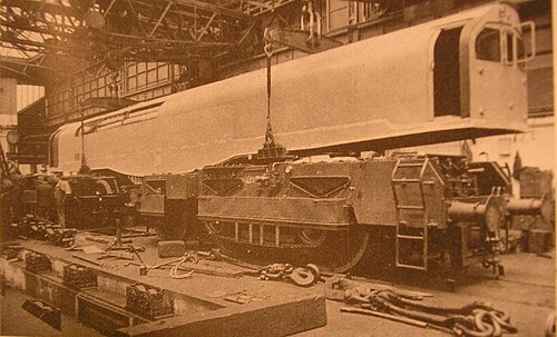 0-6-0+0-6-0 蒸気機関車の製作中の写真。機関車工場内のクレーンで、運転台とボイラーを収めた直方体の箱が 0-6-0 台車 2 台の上に降ろされている。手前にはさまざまな部品が散らばっている。