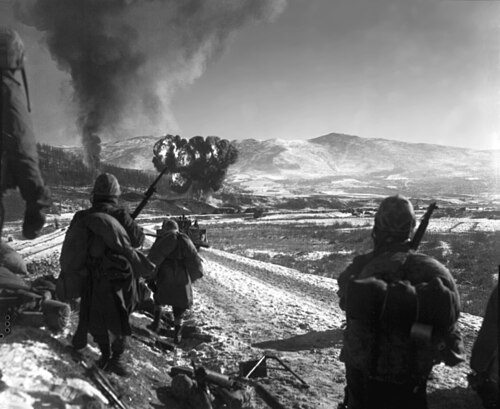 兵士たちは、飛行機が爆弾を投下するのを目の前の丘で見守っている。