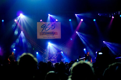 プロジェクト86がコンサートで演奏します。バンド全員が青いライトに包まれた大勢の観客の前でステージ上で演奏します。