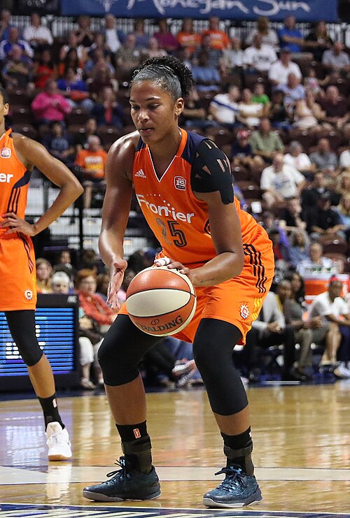 オレンジ色のユニフォームと黒いキネシオロジーテープを身に着け、フリースローラインでボールを持った選手。彼女の後ろには大勢の観客席が見える。