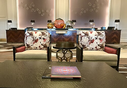 画像には、本が置かれたコーヒーテーブル、2 つの大きなアームチェア、背景の受付デスクが写っています。受付デスクの後ろの壁には大きなアート作品が飾られています。
