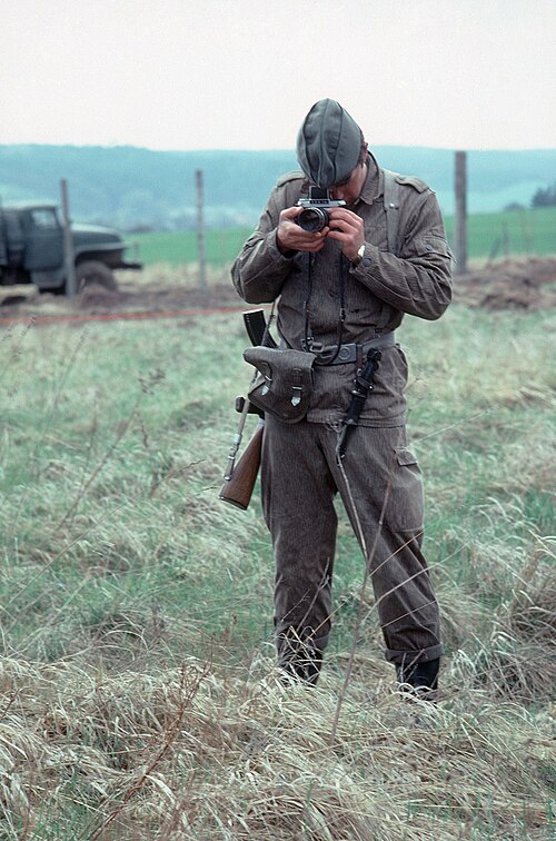 武装した東ドイツ兵が草むらに立ち、写真家を撮影している。兵士の少し後ろには、国境のフェンスとトラックが見える。