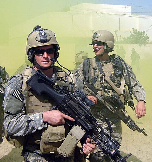米海軍特殊部隊のマイケル・モンスールは、緑色の戦闘服を着た緑色の迷彩服を着た特殊部隊の仲間と一緒。二人とも銃器を所持し、サングラスをかけている。背景には白い建物と緑色の煙が渦巻いている。