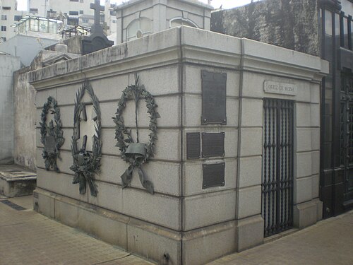 灰色の切石造りの霊廟建築の写真。外壁には金属製の花輪と銘板が取り付けられ、入り口には金属製の格子門があり、その上に四角いキューポラが載っている。