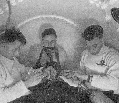 加圧室の中に3人の男性がいる。1人はマスクで呼吸しており、残りの2人は時間を計りながらメモを取っている。