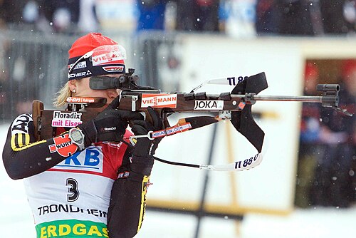 赤い帽子と背番号 3 のジャージを着た、カラフルな冬用スポーツウェアを着た女性が、ライフルを水平に構えています。ライフルの側面には広告が付いており、背景には雪が降っています。