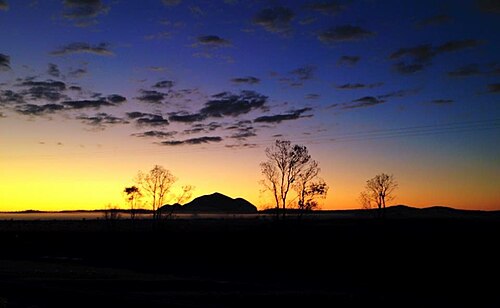 地平線上に小さな尖った山があり、前景には点在する裸木が、夕焼けの空を背景にシルエットを描いている。