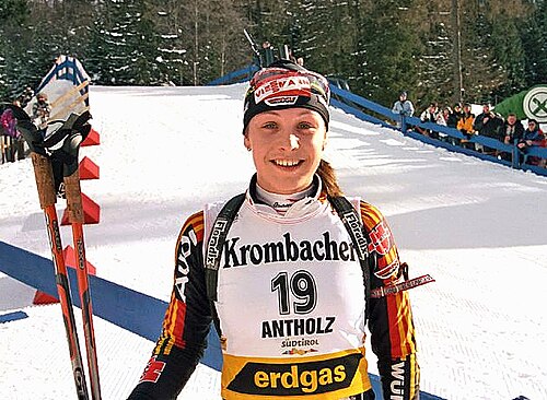 カラフルなウィンタースポーツウェアを着て、背番号19のジャージを着た若い女性が、雪に覆われた野原に立ってカメラに向かって微笑んでいる。背景には森と大勢の見物人が見える。彼女は右手にスキーのストックを持ち、背中にはライフルを背負っている。
