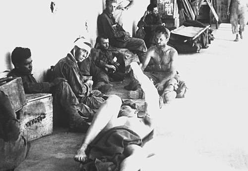 数人の男性が建物の近くに座ったり横になったりしている。包帯を巻いている人もいれば、シャツを着ていない人もいる。近くにはさまざまな箱や簡易ベッドが置かれている。