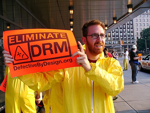 「DRM を排除せよ」というプラカードを掲げるタイベックスーツを着た男性