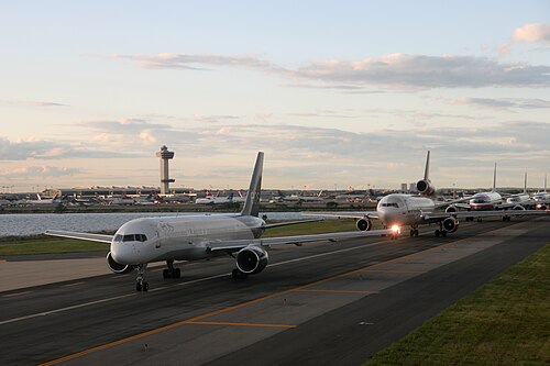5 機の大型飛行機が小さな水域の横の滑走路に一列に並んで待機しています。その向こうの遠くに空港と管制塔が見えます。