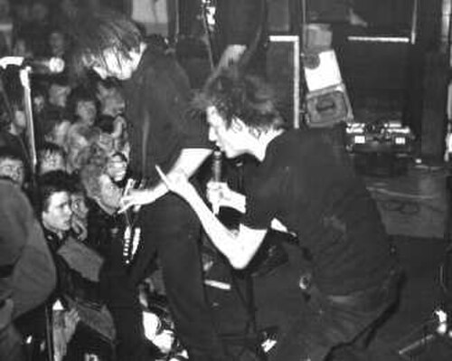 ロックバンド Crass の 2 人のメンバーが演奏している様子。左から右に、エレキギター奏者と歌手。2 人とも全身黒の服を着ています。歌手は手振りをしています。