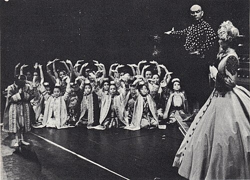 劇場の舞台の白黒写真。頭を剃り、堂々とした風格を漂わせる中年の男性が右腕を伸ばし、アジアの衣装を着た子供たちのグループを、クリノリンのドレスとボンネットを身に着けた、舞台の右手前で部分的に舞台の向こうを向いている女性に紹介している。子供たちのほとんどはひざまずいて腕を上げて挨拶している。1人の子供(おそらくチュラロンコーン皇太子)が立ち上がってお辞儀をしている。