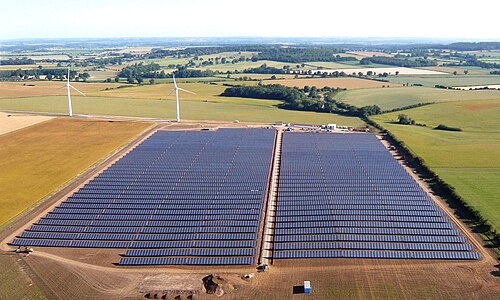 太陽光発電所の空撮写真。背景には風力発電所の一部が写っている。