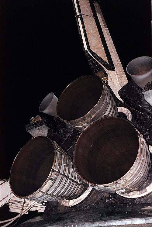 スペース シャトル オービターの尾部構造から突き出ている 3 つのベル型のロケット エンジン ノズル。クラスターは三角形に配置されており、上部に 1 つのエンジン、下部に 2 つのエンジンがあります。上部のエンジンの左右には 2 つの小さなノズルが見え、オービターの尾翼は画像の上部に向かって突き出ています。背景には夜空とパージ装置があります。