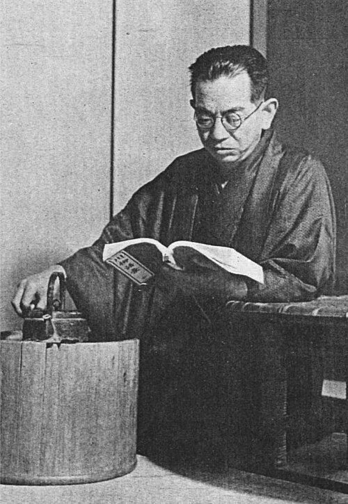 着物と眼鏡をつけた中年の日本人男性が、ひざまずいて左手に持った本を読んでいる。肘はテーブルに置き、右手はやかんに乗せている。