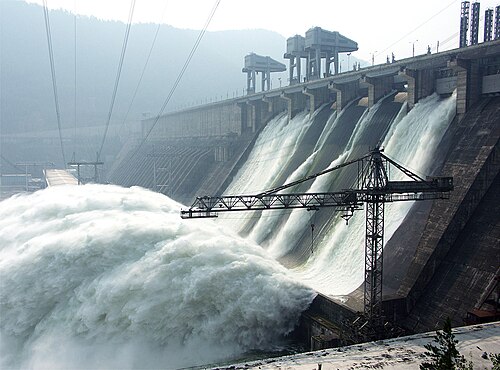 滝のように水を流す大きな貯水構造物、いくつかの電線、建設用クレーンの画像