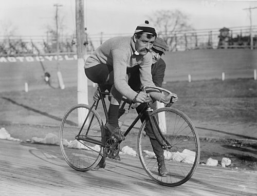 古い自転車に乗った男性の白黒写真。もう一人の男性が自転車を抱えたり押したりしています。