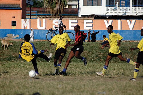 サッカーをしている5人の男性。1人は赤と黒の縞模様のジャージを着ており、4人は黄色のジャージを着ている。