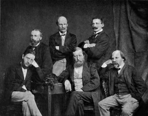黒いジャケットとネクタイを締めた中年男性6人のグループ。3人が座り、3人が立っている。