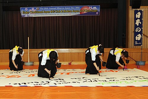 袴を着た日本の女子高校生4人が、床に固定された幅5メートル、高さ3メートルの大きな紙に書道をしている。