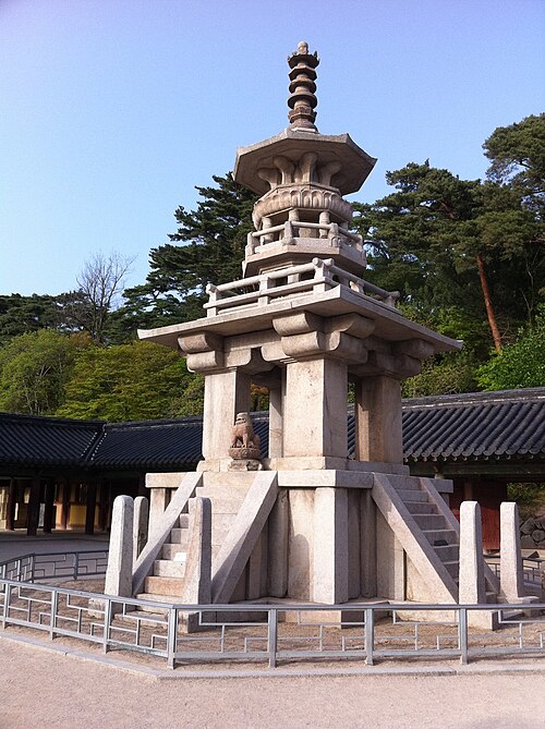 精巧な段、小さな獅子像、階段のある石塔。背景には青い空と建物の屋根、木々が描かれている。