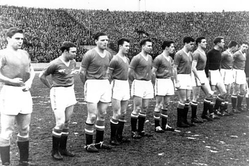試合前に整列するサッカーチームの白黒写真。昔ながらのサッカーのユニフォームを着た 11 人の男性が一列に並んでいる。10 人は濃い色のシャツ、白いショートパンツ、黒いソックスを履いており、もう 1 人はさらに濃い色のシャツを着ている。選手たちの後ろには、東ヨーロッパ風のサッカー スタジアムの巨大なオープン スタンドが 1 つ見え、観客でいっぱいになっている。