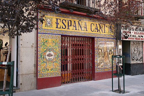 塗装されたセラミックタイルで覆われた閉店したバーのファサード。上部には「14」と「ESPAÑA CAÑÍ」と書かれています。柵で囲まれた門の左側には、装飾されたパネルに闘牛士の帽子があります。門の右側には、裸の女性、アンダルシアの衣装を着た2人の恋人、その他の人物が描かれています。