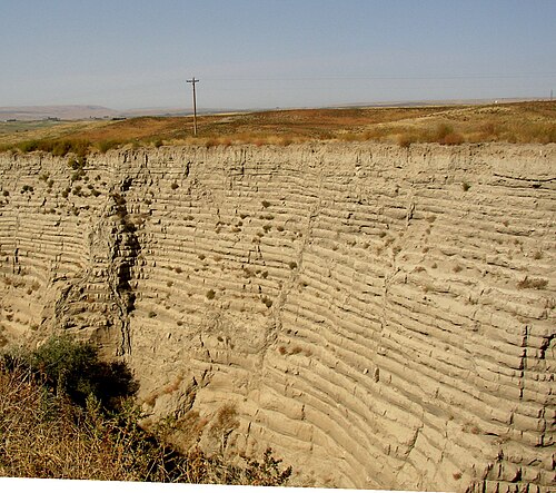 この写真は、周囲の平らな土壌に切り込まれた別の峡谷を示しています。この峡谷には約 30 の水平堆積層があり、それぞれの層は下の層と明確に区​​別されています。峡谷の上には遠くに電柱が見えます。この電柱のおかげで、切り込みの深さが 30 ～ 40 フィート (9.1 ～ 12.2 メートル) であることが分かります。手前には峡谷の近くの縁が見えます。このことから、峡谷がかなり狭く、壁が急峻であることが分かります。