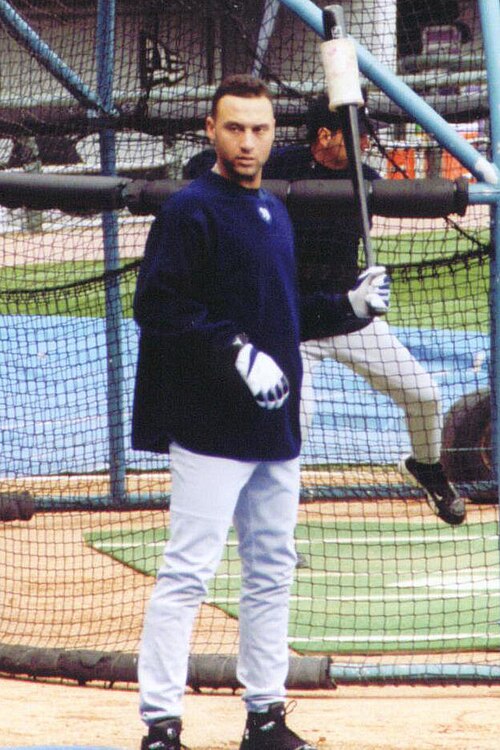 デレク・ジーターは野球ユニフォームの上にネイビーの長袖シャツを着て、左手に重り付きのバットを持ち、バッティングケージの前に立っています。