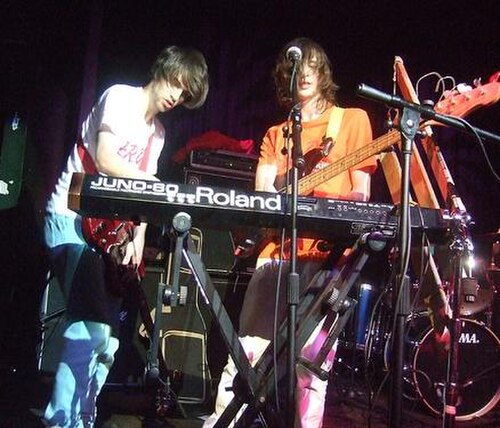ステージにはエレキギター奏者とベーシストがいて、その横には「JUNO-60 Roland」シンセサイザーキーボードがあり、ギタリストがキーボードを演奏しています。背景にはドラムキットがあります。