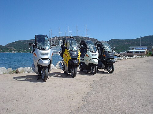 湖の前に4台のBMW C1スクーターが並んで駐車されており、背景には山々と大型船のマストが見える。