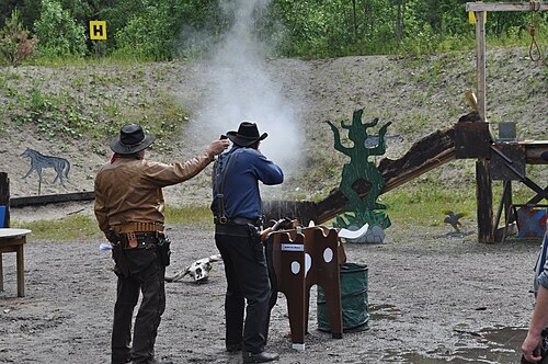 カウボーイ アクション シューターがレバー アクション ライフルを鋼鉄の標的に向けて発射しています。左側の射撃場責任者は時間を計測するために射撃タイマーを持っています。