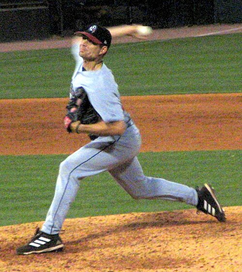 灰色の野球ユニフォームと黒い帽子をかぶった男性が、投手マウンドに立って右手で野球ボールを投げる準備をしている。