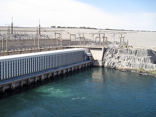 穏やかな湖のそばに建つ大きなコンクリートの建物。建物のすぐそばには、多数の大きな送電塔がそびえ立っている。