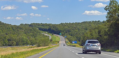中央分離帯のある高速道路。右側にカメラがあり、短い下り坂を進んでいます。前方右側に銀色の車があり、遠くに近くの出口とそこにあるサービスを示す緑と青の標識があります。
