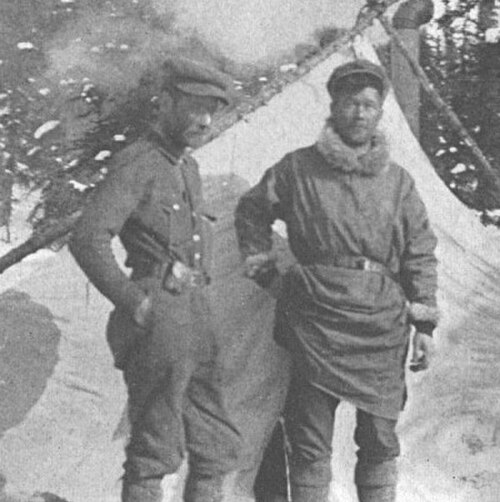 モノクロ写真には、テントと雪に覆われた常緑樹の前に立つ二人の男性が写っている。