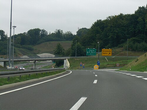 高速道路が典型的な出口と高架道路に近づいており、片持ち梁式ガントリーに設置された方向標識が道路の片側から見える。