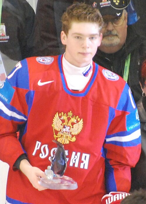 赤いロシアのアイスホッケーユニフォームを着た、短くて明るい茶色の髪の白人男性が、小さなガラスの賞状を持っています。