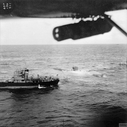 波立つ海に浮上した潜水艦の航空写真。小型船が近くにあり、両方とも静止しているように見える。飛行機の左翼の一部が見え、そこに爆弾がぶら下がっている。