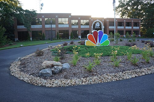WYFF のロゴが入った 2 階建てのオフィス ビル。正面には、立体的な NBC の孔雀をあしらった大きな造園要素を備えた待合室と私道があります。