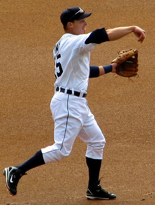 白いユニフォームを着た野球選手が、投球動作のフォロースルーの姿勢をとっている。腕は前に伸ばされ、手は下を向いており、左手にはグローブがはめられている。左足は地面にしっかりと着地し、右足は後ろに浮かせている。