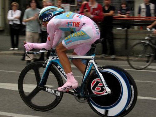 ピンク色の服と靴を身に着けた男性が、自転車に身をかがめて座っている。背景には人々が見ている。