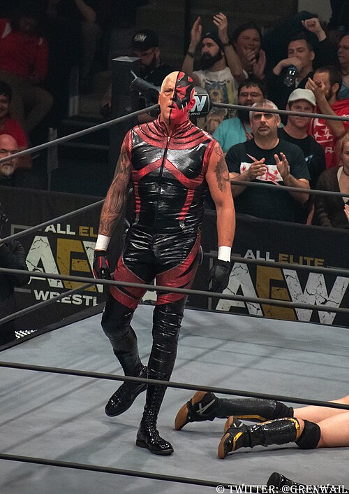 赤い衣装を着て、顔の半分を赤く塗った男性がプロレスリングのリングにいるカラー写真