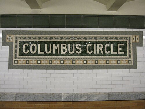 IRT プラットフォームにあるモザイクの銘板には、オレンジ色のタイルで囲まれた大文字の「Columbus Circle」の文字があります。銘板の下には白いタイル張りの壁があります。