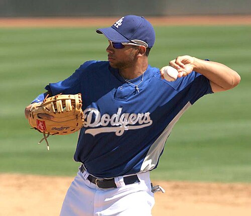 胸に「DODGERS」と書かれた青い野球ユニフォームを着て、右手に野球グローブをはめた男性が、左手で野球ボールを投げる準備をしている。