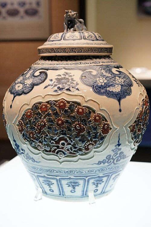 元代の青白磁で、青と赤の釉薬で透かし彫りの花模様（蓮の花びら、草、牡丹）が描かれ、蓋には獅子の飾りが付けられている。
